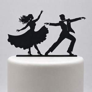Peut inclure: Une décoration de gâteau en silhouette noire représentant un couple dans une pose de danse de salon. La silhouette de gauche porte une robe fluide avec de longs cheveux, tandis que celle de droite porte un costume formel avec un nœud papillon.