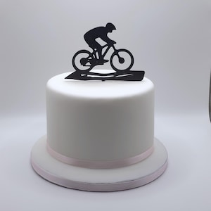 May include: A round white cake with a smooth fondant finish and a light pink ribbon border at the base. A black silhouette cake topper featuring a cyclist riding a mountain bike is positioned on the top center of the cake.