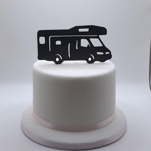 May include: A black campervan cake topper sits atop a white cake with a pink ribbon. The campervan is a silhouette with details like windows and wheels. The cake is on a white cake stand.