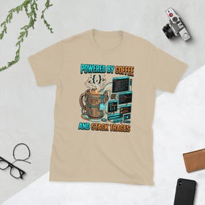 May include: A beige t-shirt featuring a graphic of a coffee mug connected to computer code windows. The text reads POWERED BY COFFEE AND STACK TRACES. The mug is labeled CAFFEINE TYPE: CF-3 and displays programming symbols like curly braces.