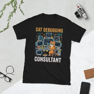 May include: A black t-shirt featuring a graphic of an orange cat wearing glasses and holding a magnifying glass while sitting at a computer desk. The text reads CAT DEBUGGING CONSULTANT, surrounded by computer monitors, code, and small yellow ducks.