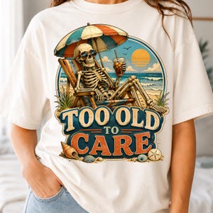 May include: An off-white t-shirt featuring a graphic of a skeleton wearing sunglasses, sitting in a beach chair under an umbrella, holding a drink. The text reads TOO OLD TO CARE, with illustrations of seashells and a sunset over the ocean.
