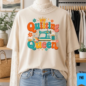 May include: A beige t-shirt featuring a graphic print with the text Quilting Queen in orange and teal retro-style lettering. The design includes a teal sewing machine, a spool of thread, a patchwork quilt square, crowns, and colorful flowers.