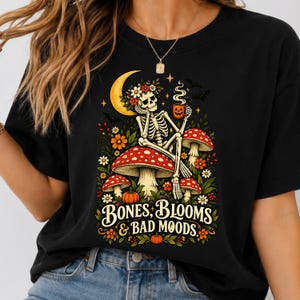 May include: A black t-shirt featuring a graphic illustration of a skeleton sitting on a large red and white spotted mushroom. The skeleton wears a flower crown, holds a jack-o'-lantern mug, and is surrounded by foliage, pumpkins, and a crescent moon. The text reads BONES, BLOOMS & BAD MOODS.