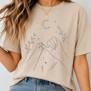 May include: A beige short-sleeve t-shirt featuring a black line art graphic of a human hand and a cat paw performing a pinky promise. The design includes a crescent moon, small stars, and delicate leafy branches surrounding the central figures.