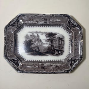 Può includere: Un piatto ottagonale in ceramica antica con una scena di paesaggio in bianco e nero. Il piatto ha un bordo decorativo con motivi floreali e geometrici. L'immagine centrale raffigura un paesaggio classico con edifici, alberi e figure.