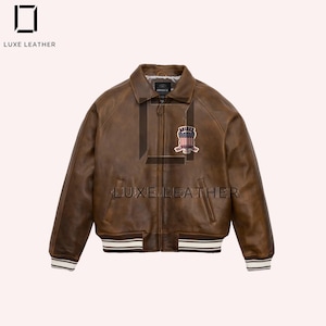 Puede incluir: Chaqueta bomber de cuero marr&oacute;n con cierre de cremallera, cuello y parche en el pecho. La chaqueta tiene pu&ntilde;os y dobladillo a rayas blancas y marrones. La marca "LUXE LEATHER" est&aacute; impresa en la chaqueta.