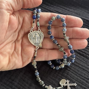 May include: A rosary with blue and gray beads and silver-toned metal accents. The centerpiece features an image of the Virgin Mary. The rosary also includes a cross pendant with the words "MADE WITH LOVE".