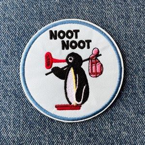May include: Circular embroidered patch with a cartoon penguin carrying a red horn and a bag, with the text "NOOT NOOT". The patch is on a denim background. The patch has a light blue border.