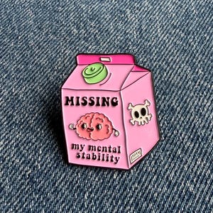 May include: A pink enamel pin shaped like a milk carton featuring a cartoon brain, a skull, and the text MISSING my mental stability. The pin has a green cap detail and black outlines, presented as a graphic accessory with a humorous design.