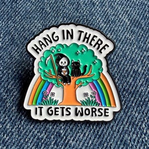 May include: A metal enamel pin featuring a grim reaper and a black cat sitting in a green tree under a rainbow. The text HANG IN THERE is arched above the tree, and the text IT GETS WORSE is printed in black at the bottom on a white background.