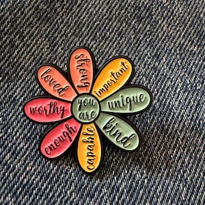 May include: A flower-shaped enamel pin with a black outline. The centre circle reads "you are" and the eight petals feature the words "strong," "important," "unique," "kind," "capable," "enough," "worthy," and "loved" in black cursive script.