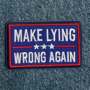 May include: A rectangular embroidered patch with a blue background, a red border, and white text that reads "MAKE LYING WRONG AGAIN" with three white stars centred between the two lines of text, all set against a blue denim fabric background.