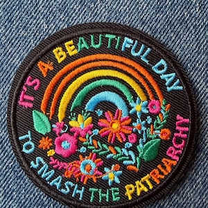 May include: A circular black embroidered patch featuring a colourful rainbow and various flowers in shades of pink, yellow, orange, and blue. The text "IT'S A BEAUTIFUL DAY TO SMASH THE PATRIARCHY" is stitched in a circular pattern around the design.