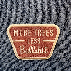 May include: A trapezoidal embroidered patch with a rust-coloured background and a cream-coloured border. The text reads MORE TREES LESS Bullshit in cream-coloured thread, with a horizontal line separating the words TREES and LESS.