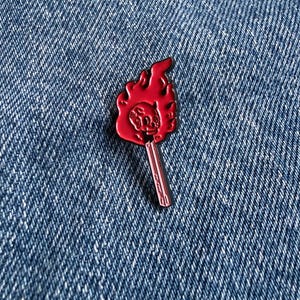 May include: A metal enamel pin shaped like a lit matchstick. The head of the match is a vibrant red flame with a small, embossed skull design centred within the fire. The matchstick body is a light pinkish-red colour with thin, dark outlines.