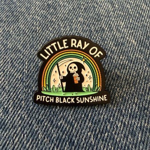 May include: A black enamel pin featuring a cartoon grim reaper holding a scythe and an orange drink. A colourful rainbow arches over the figure against a light background with small stars. The text reads LITTLE RAY OF PITCH BLACK SUNSHINE.