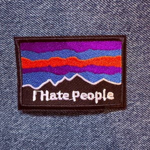 May include: A rectangular embroidered patch featuring a mountain landscape design with horizontal bands of purple, blue, and orange thread above a black base. The text "I Hate People" is stitched in white lettering across the bottom black section.