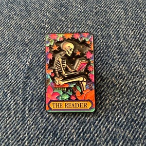 May include: A rectangular enamel pin featuring a skeleton sitting and reading a book. The design is surrounded by colourful flowers and stars against a dark background. The text THE READER is printed in a yellow banner at the bottom of the pin.