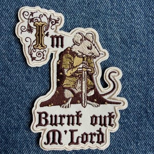 May include: An embroidered patch featuring a mouse dressed in medieval armour and a dark brown cloak, holding a sword. The text reads "I'm Burnt out M'Lord" in a gothic font. The design includes decorative scrollwork and a cream-coloured border.