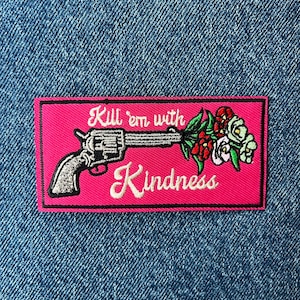 May include: A rectangular embroidered patch with a hot pink background featuring a silver revolver with a bouquet of red, white, and green flowers emerging from the barrel. The text "Kill 'em with Kindness" is stitched in white cursive script.
