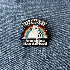 May include: A black and white pin featuring a raccoon sitting on a rainbow. The pin has the text "Your Little Ray of Sarcastic Sunshine Has Arrived" in white lettering.