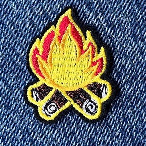 May include: Embroidered patch with a campfire design. The patch has a black border, with a yellow flame outlined in red. The logs are brown with white details. The patch is on a blue denim background.