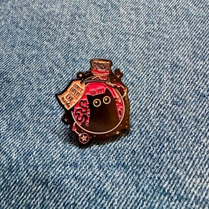 May include: A metal enamel pin shaped like a potion bottle containing a black cat with wide eyes. The bottle features a pink liquid, a cork stopper, and a hanging tag that reads DON'T OPEN EVIL inside, with the text DO IT! printed above the cat.