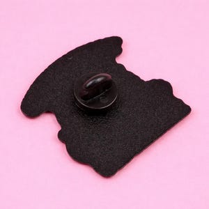Op de afbeelding: Een zwarte emaille pin met een afgeronde vorm en een pin back. De pin staat tegen een roze achtergrond. De pin is waarschijnlijk een decoratief accessoire.