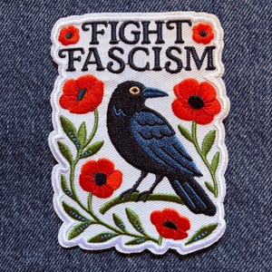 May include: A rectangular embroidered patch with a white background featuring a black crow perched on a green branch surrounded by red poppy flowers and green leaves. The text FIGHT FASCISM is embroidered in black capital letters at the top.