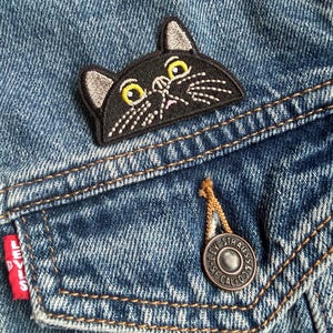 May include: A black cat embroidered patch featuring yellow eyes, white whiskers, and grey inner ears, attached to a blue denim jacket pocket. The pocket features a copper-coloured metal button embossed with the text Levi Strauss & Co.