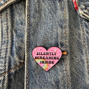 May include: A heart-shaped enamel pin with a pink background, featuring the black text "SILENTLY SCREAMING INSIDE" in the centre. The design includes a small rainbow at the bottom, tiny white hearts, and star accents, all outlined in black metal.