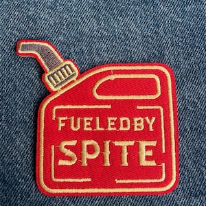 May include: A red embroidered patch shaped like a fuel can with a grey spout. The patch features gold-coloured stitching that outlines the can and spells out the text FUELED BY SPITE in a bold, block-style font centred on the front of the container.