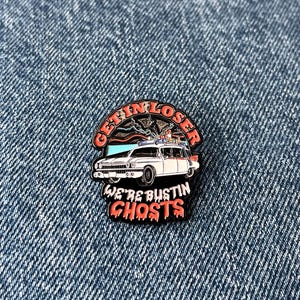 May include: Enamel pin depicting the Ecto-1 from Ghostbusters. The pin reads "GET IN LOSER" above the car and "WE'RE BUSTIN' GHOSTS" below. The car is white with red and blue details, set against a blue denim background.