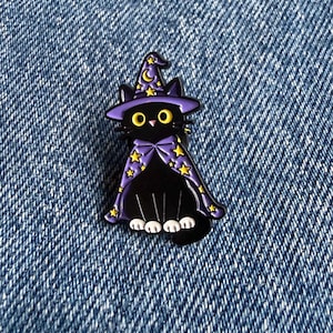 May include: A black cat enamel pin wearing a purple wizard hat and a matching purple cape, both decorated with yellow stars and a crescent moon. The cat has large yellow eyes, a small pink nose, and white paws, rendered in a cartoon style.