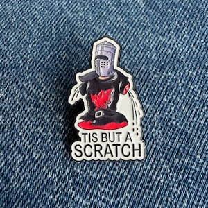 May include: A metal enamel pin featuring a cartoon knight wearing a grey helmet and black tunic with missing arms, standing in a red puddle. The design includes white liquid spray effects and the black text TIS BUT A SCRATCH at the bottom.
