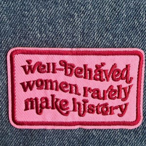 May include: A rectangular embroidered patch with a light pink background and a dark pink border. The patch features the dark pink embroidered text "well-behaved women rarely make history" in a stylised, cursive-inspired font, centred on the fabric.