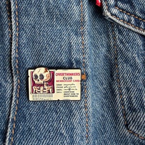 May include: A rectangular enamel pin featuring a cartoon skull and the text OVERTHINKERS CLUB MEMBERSHIP CARD. The card includes fields for name, phone, email, and skill, with the text OVERTHINK ABOUT HOW I'M OVERTHINKING. The pin is attached to blue denim.