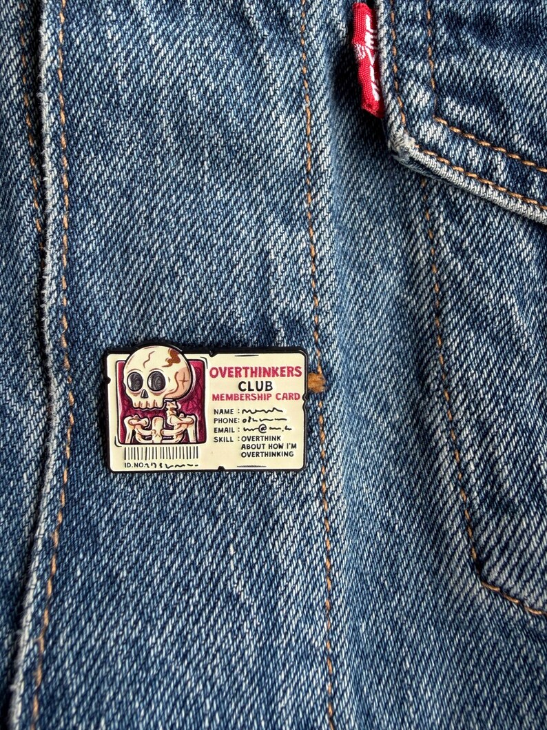 May include: A rectangular enamel pin featuring a cartoon skull and the text OVERTHINKERS CLUB MEMBERSHIP CARD. The card includes fields for name, phone, email, and skill, with the text OVERTHINK ABOUT HOW I'M OVERTHINKING. The pin is attached to blue denim.