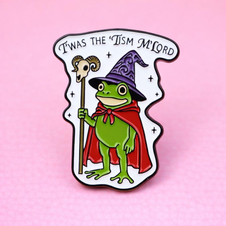 May include: Enamel pin featuring a green frog dressed as a wizard, wearing a purple hat and red cape. The frog holds a staff with a skull on top. The text "Twas the 'Tism M'Lord" is above the frog.