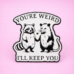 Op de afbeelding: Zwart-witte pin met een wasbeer en een opossum die elkaars hand vasthouden, met de tekst "YOU'RE WEIRD I'LL KEEP YOU" in een zwarte rand. De achtergrond is roze.