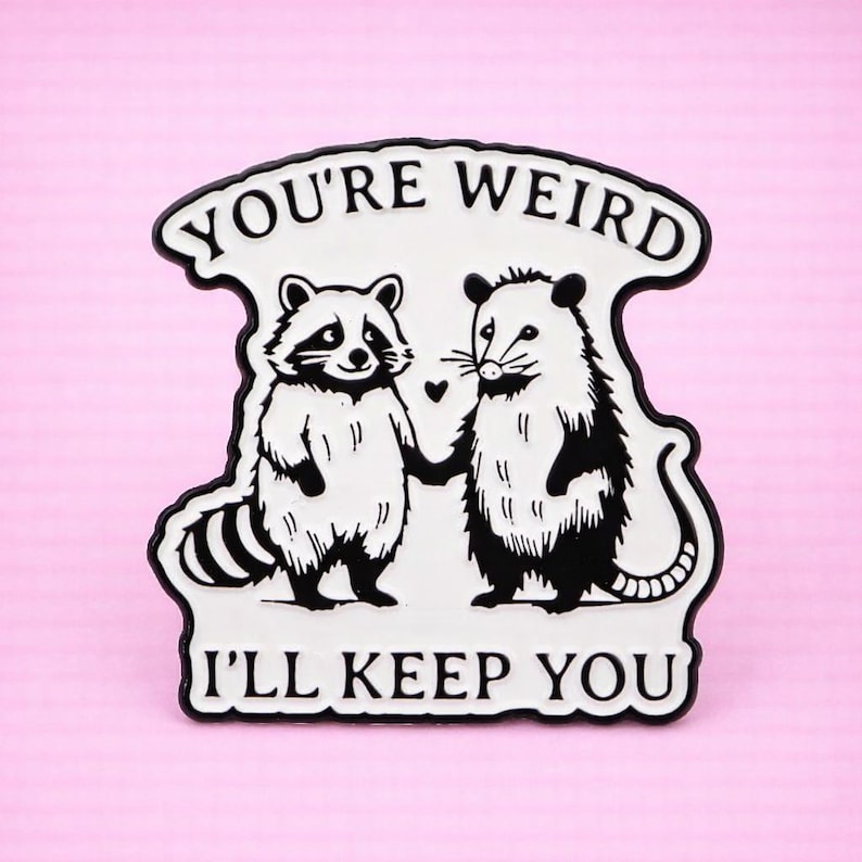 Op de afbeelding: Zwart-witte pin met een wasbeer en een opossum die elkaars hand vasthouden, met de tekst "YOU'RE WEIRD I'LL KEEP YOU" in een zwarte rand. De achtergrond is roze.