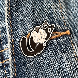 May include: A black enamel pin depicting a cartoon cat clutching a white D20 die. The cat has large eyes and a surprised expression. The pin is set against a blue denim background.