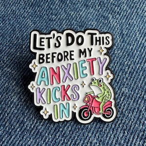 May include: A white enamel pin with black outlines featuring a green frog riding a pink scooter. The text reads LET'S DO THIS BEFORE MY ANXIETY KICKS IN in black, blue, purple, and pink lettering, accented by small gold-coloured star shapes.