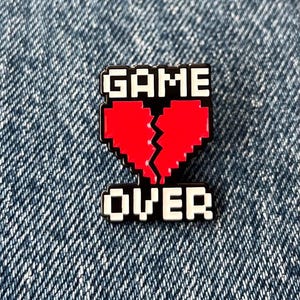 May include: A black and red enamel pin with the words "GAME OVER" in white pixelated font. The pin features a broken red heart in the centre, set against a black background. The pin is on a blue denim background.
