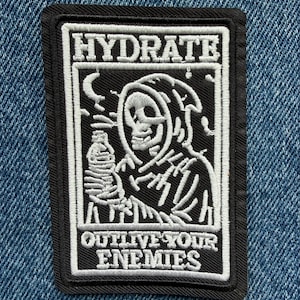May include: A rectangular embroidered patch with a black background and white stitching. It features a hooded skeleton holding a water bottle, a crescent moon, and the text HYDRATE at the top and OUTLIVE YOUR ENEMIES at the bottom of the design.