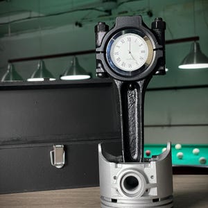 May include: A unique clock made from automotive parts. The clock face is set within a black connecting rod, with a silver piston base. The clock has a white face with black Roman numerals and black hands. A black box is in the background.