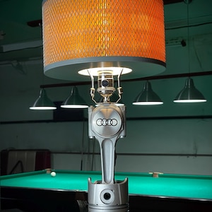 Może przedstawiać: Unikalna lampa stołowa z pomarańczowym kloszem z siatki i srebrnym tłokiem jako podstawą. Lampa ma logo Audi i stoi na czarnej okrągłej podstawie. W tle widać stół bilardowy.