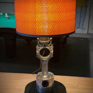 May include: A unique table lamp with an orange, diamond-patterned lampshade. The base is made from a repurposed engine piston and connecting rod, mounted on a dark wooden base. The lamp emits a warm, inviting glow.