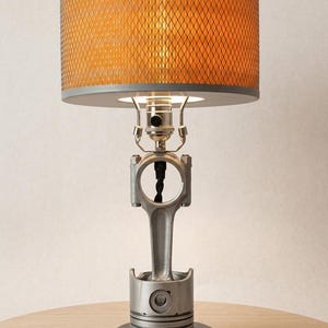 Puede incluir: Una lámpara de mesa con un pistón de motor y una biela de metal plateado como base, montada sobre un soporte circular negro. La lámpara tiene una pantalla cilíndrica con un exterior de malla metálica y un forro interior de tono naranja.